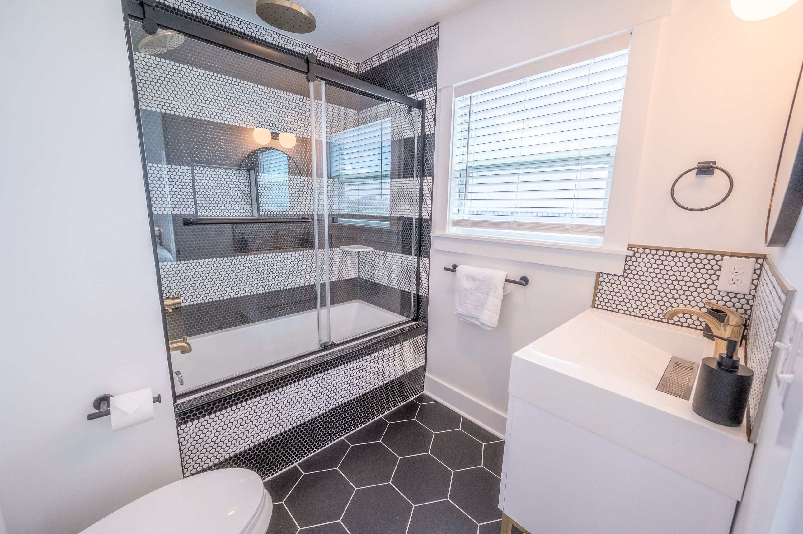 A modern bathroom with a glass-enclosed shower, a white sink with a black soap dispenser, and a hexagonal tile floor.