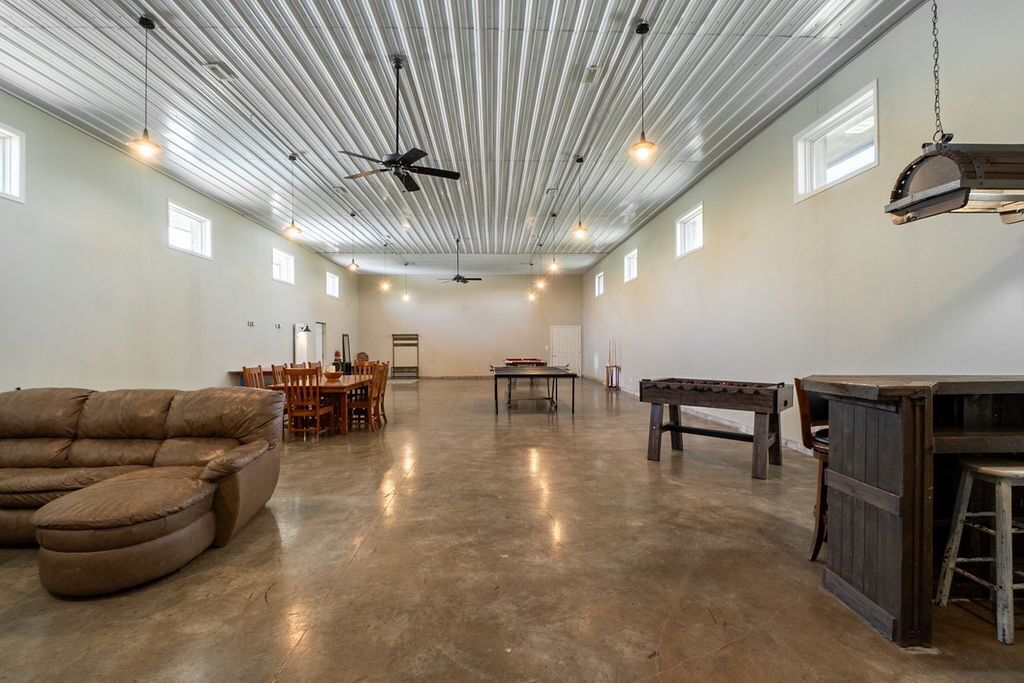 A spacious room with a high ceiling, exposed beams, and a ceiling fan. There is a wooden table and chairs in the center, and a sofa on the left.