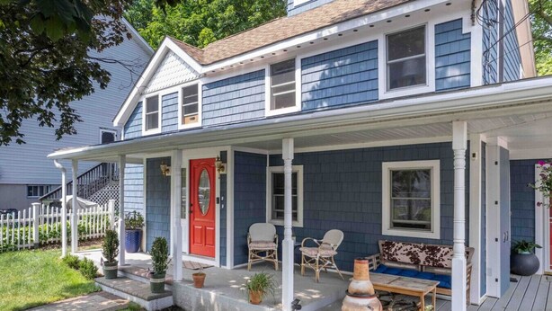 A two-story house with a porch, a red door, and a small outdoor seating area with chairs and a table.