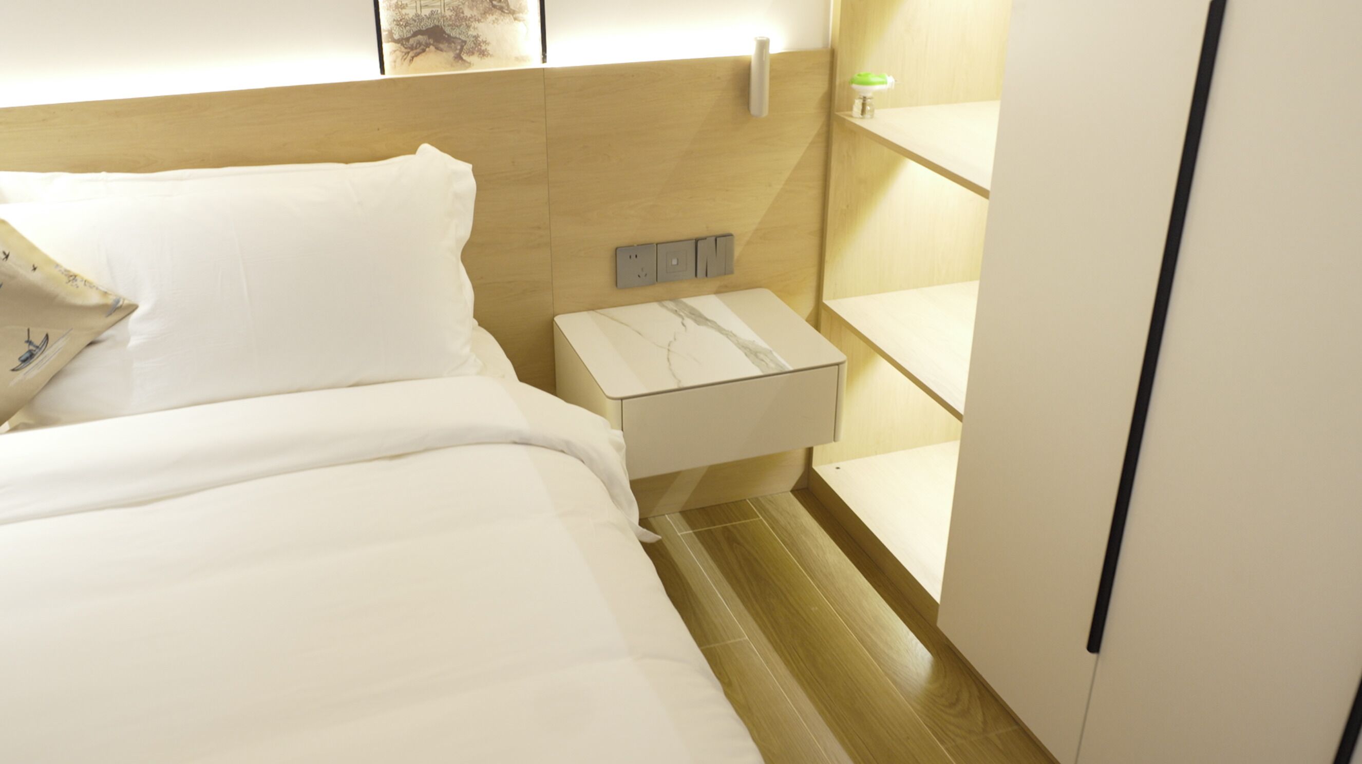 A hotel room with a bed, a nightstand, a wall-mounted light, and a shelf.