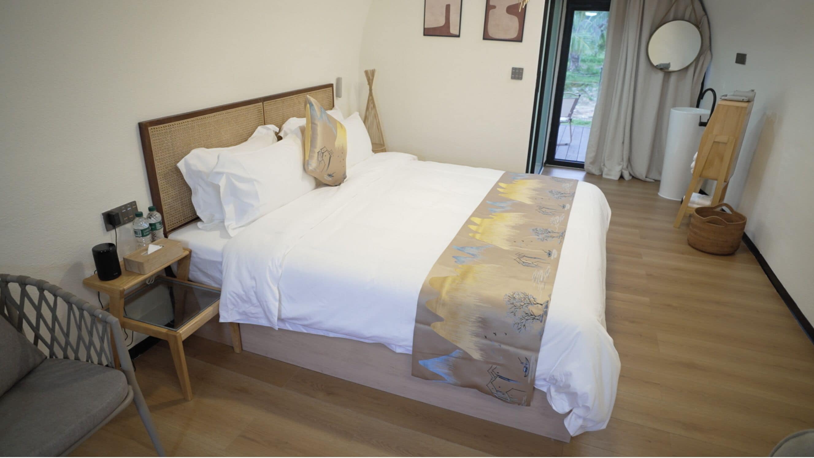 A neatly made bed with white linens and a patterned throw blanket, a bedside table with a lamp and water bottles, a chair, and a view of the outdoors through a glass door.