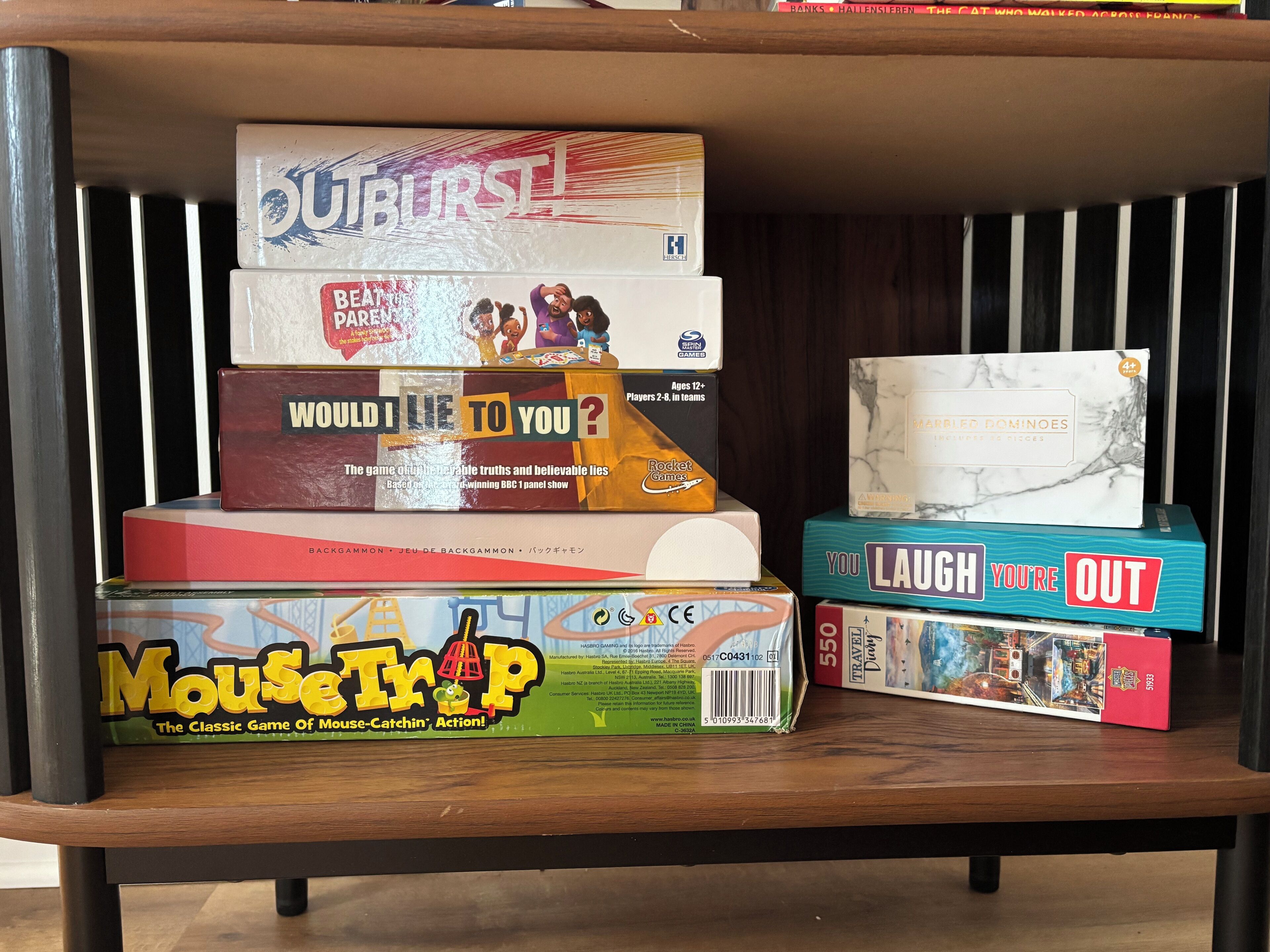 Un estante con varios juegos de mesa, incluyendo 'Would I Lie to You?', 'Mouse Trap', 'You Laugh You're Out' y 'Busted!'. 