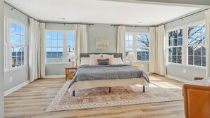 A spacious bedroom with a large bed, two bedside tables, and a view of the outdoors through large windows. - Kick back and enjoy the Chesapeake & this 3-bedroom, relaxing, stylish space. (Prince Frederick)