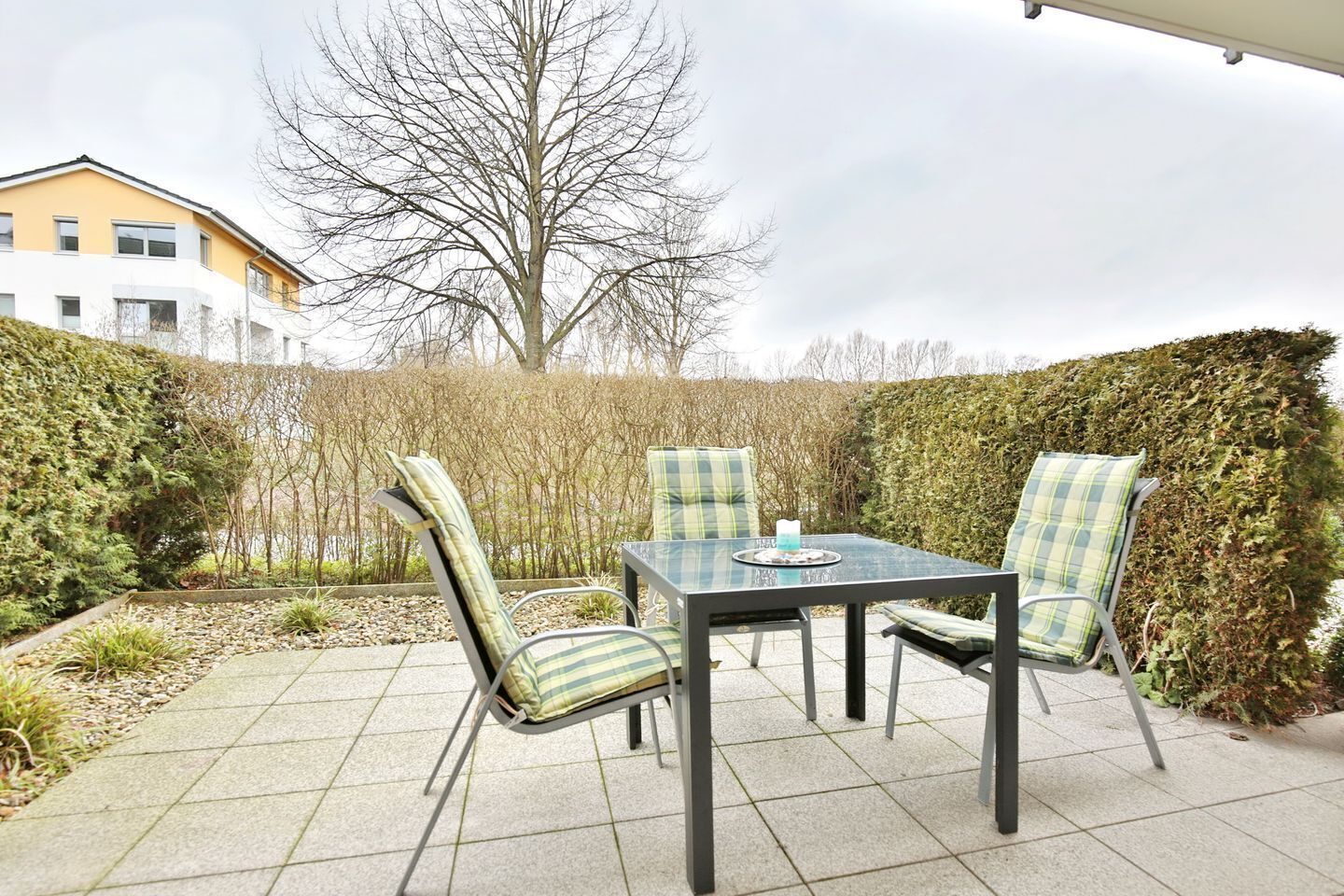 An outdoor patio with a table and chairs, surrounded by hedges and a building in the background.