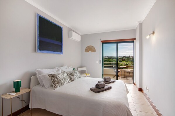 Une chambre à coucher comprenant un lit, une table de chevet, une lampe, une vue sur l’extérieur par une porte vitrée et un climatiseur fixé au mur.
