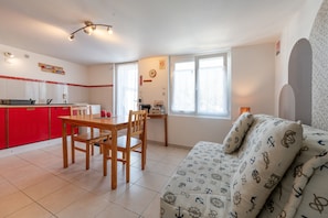 Een ruimtelijk ingerichte leefruimte met een eethoek, kitchenette en een slaapbank met nautische beddengoed.