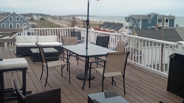 Une terrasse avec une table et des chaises, un parasol rouge et une vue sur les maisons et l’océan.