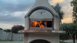 A stone pizza oven with a fire burning inside, surrounded by a wooden deck and a fenced yard.
