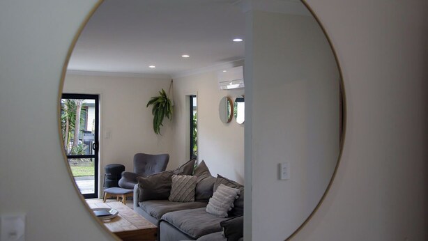 A modern living room with a grey sofa, a small table, and a chair. There is a plant on the wall and a window with a view of greenery outside.