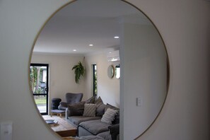 Ein modernes Wohnzimmer mit einem grauen Sofa, einem kleinen Tisch und einem Sessel. An der Wand steht eine Pflanze und das Fenster bietet Blick ins Grüne.