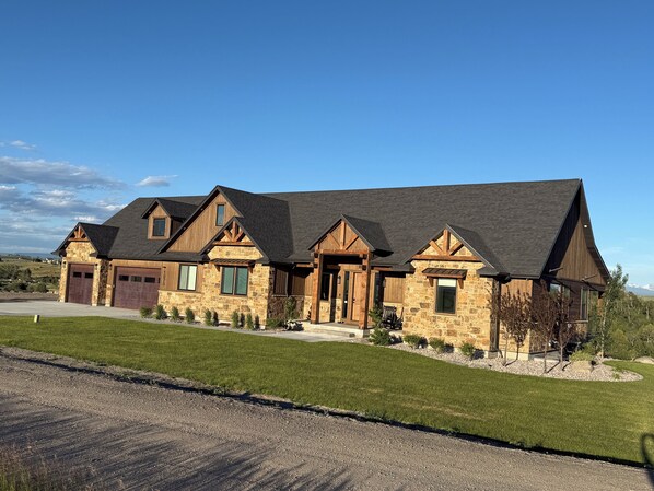 A large, multi-story house with a stone facade, multiple garage doors, and a well-maintained lawn. - River front w/ view of Tetons - Fall River Vista (Ashton)