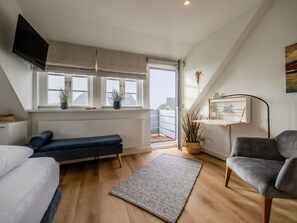 Een moderne slaapkamer met een bed, een bankje, een bureau, een stoel, een televisie en een raam met planten.