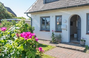 Een wit huis met een grijs dak, een duidelijke '3' op de muur, en een kleine tuin met roze bloemen.