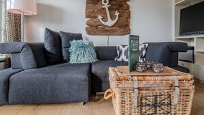 A modern living room with a grey sofa, a blue fluffy pillow, a wicker coffee table with decorative items, and a wooden wall decoration. - House Dünenwall, App. 1 - Part of the house near the beach with approx. 80 m² and 3 rooms, for (Wenningstedt-Braderup (Sylt))