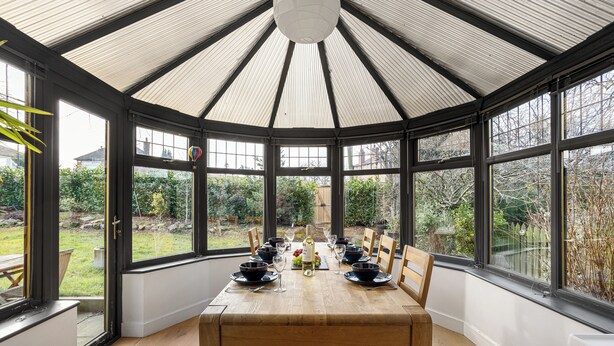 Un invernadero con una mesa de madera dispuesta para comer, ventanas amplias y vistas al jardín.