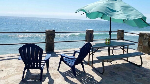 A seaside patio with two chairs and a table under a large umbrella, overlooking the ocean.