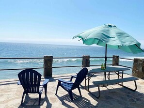 Une terrasse en bord de mer avec deux chaises et une table, protégée par un grand parasol, d’où l’on a une vue sur l’océan.