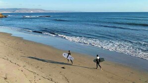 A beach with two people walking with surfboards, a clear sky, and calm sea.