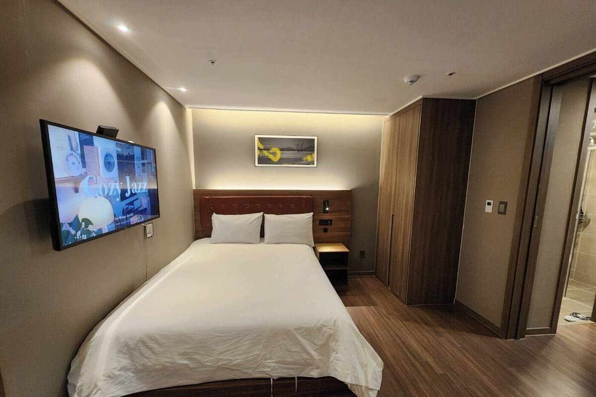 Quarto de hotel moderno com uma cama grande, televisão de tela plana na parede, cabeceira de madeira e uma mesinha de cabeceira.