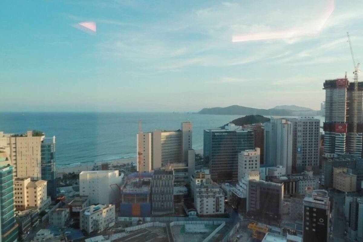 Paisagem costeira com edifícios altos, vista para o mar e uma cadeia de montanhas ao longe.