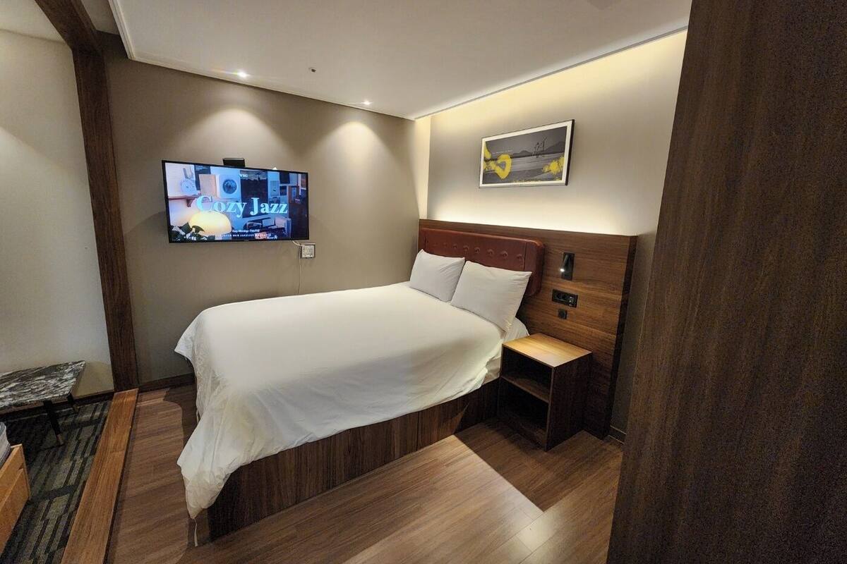 Quarto de hotel moderno com uma cama grande, uma televisão presa na parede e uma pequena mesa de cabeceira.