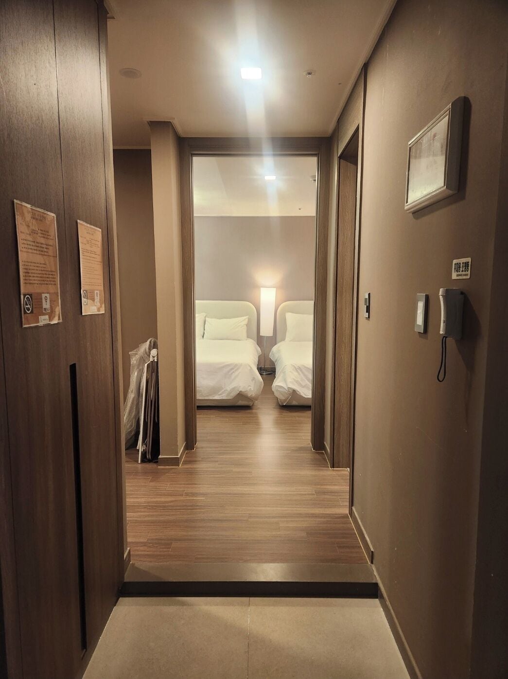 Pasillo de hotel que conduce a una habitación con dos camas, suelo de madera y entrada alfombrada.