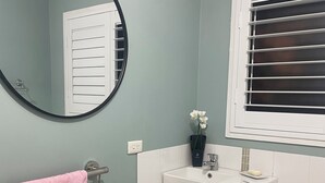 Une salle de bain avec un miroir rond, un lavabo, une fenêtre avec des stores et une serviette rose sur un porte-serviettes.