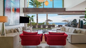 Uma sala de estar moderna com uma grande janela com vista para o oceano, mobiliada com cadeiras vermelhas e um sofá branco, e com uma televisão de tela plana.