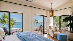 Une chambre avec un grand lit, une vue sur la mer, un balcon avec un fauteuil et un luminaire suspendu.