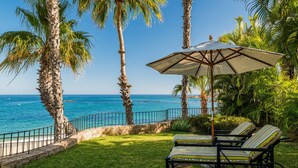 Une terrasse dotée de deux chaises longues à rayures et d’un grand parasol, offrant une vue imprenable sur la côte, avec des palmiers et l’océan.