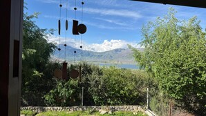 Blick durch ein Fenster, mit einer hängenden Windglocke, einer Steinmauer und einer Landschaft mit Bäumen und Bergen im Hintergrund.