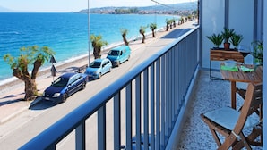Un balcón con vista al mar, una mesa de madera con sillas, y coches aparcados a lo largo de la calle.