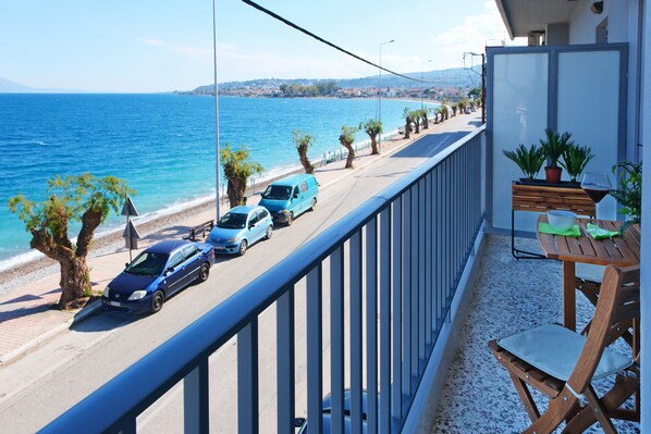 A balcony with a view of the sea, a wooden table with chairs, and parked cars along the street.