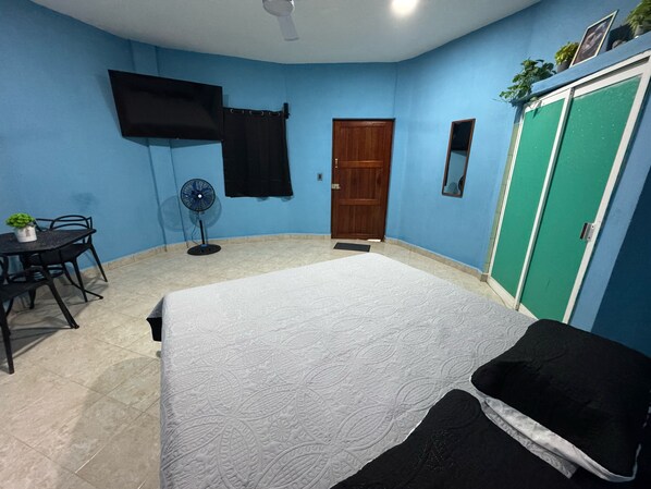 A bedroom with a bed, a desk with a chair, a standing fan, a wooden door, and a green wardrobe. - Hotel Bambús (Felipe Carrillo Puerto)