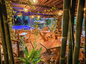 An outdoor dining area with wooden tables and chairs, surrounded by bamboo plants and hanging lights. - Hotel Bambús (Felipe Carrillo Puerto)