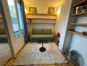 Un salon chaleureux avec un canapé vert, une petite table, une lampe rouge et un ventilateur. Il y a des étagères au-dessus du canapé et une fenêtre avec des rideaux.