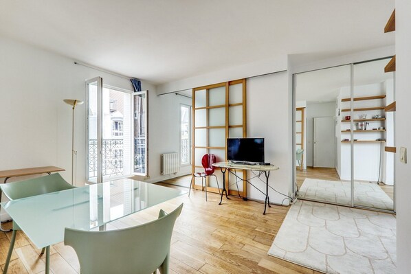 Ein modernes Wohnzimmer mit einem Glastisch, zwei Stühlen, einem Fernseher auf einem Schrank und Blick auf die Stadt durch große Fenster.