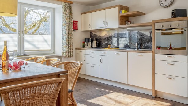 A modern kitchen with white cabinets, a wooden countertop, and a built-in oven. There is a dining area with wicker chairs and a table, and a window with curtains. - Large holiday apartment, 200m to the ski bus (Fieberbrunn)
