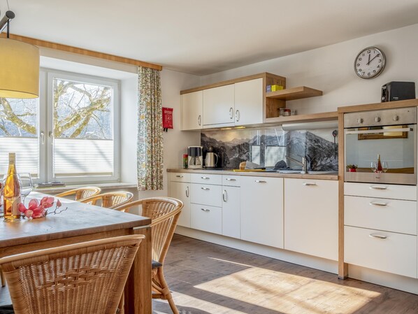 A modern kitchen with white cabinets, a wooden countertop, and a built-in oven. There is a dining area with wicker chairs and a table, and a window with curtains.