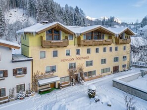 Un immeuble à plusieurs étages doté de balcons, portant l’inscription « Pension Dorfparter », entouré d’arbres et de montagnes couverts de neige.