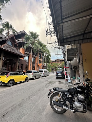 A street with parked cars and a motorcycle, buildings with balconies and palm trees, and overhead electrical wires. - Tnon Samui Hostel (Koh Samui)