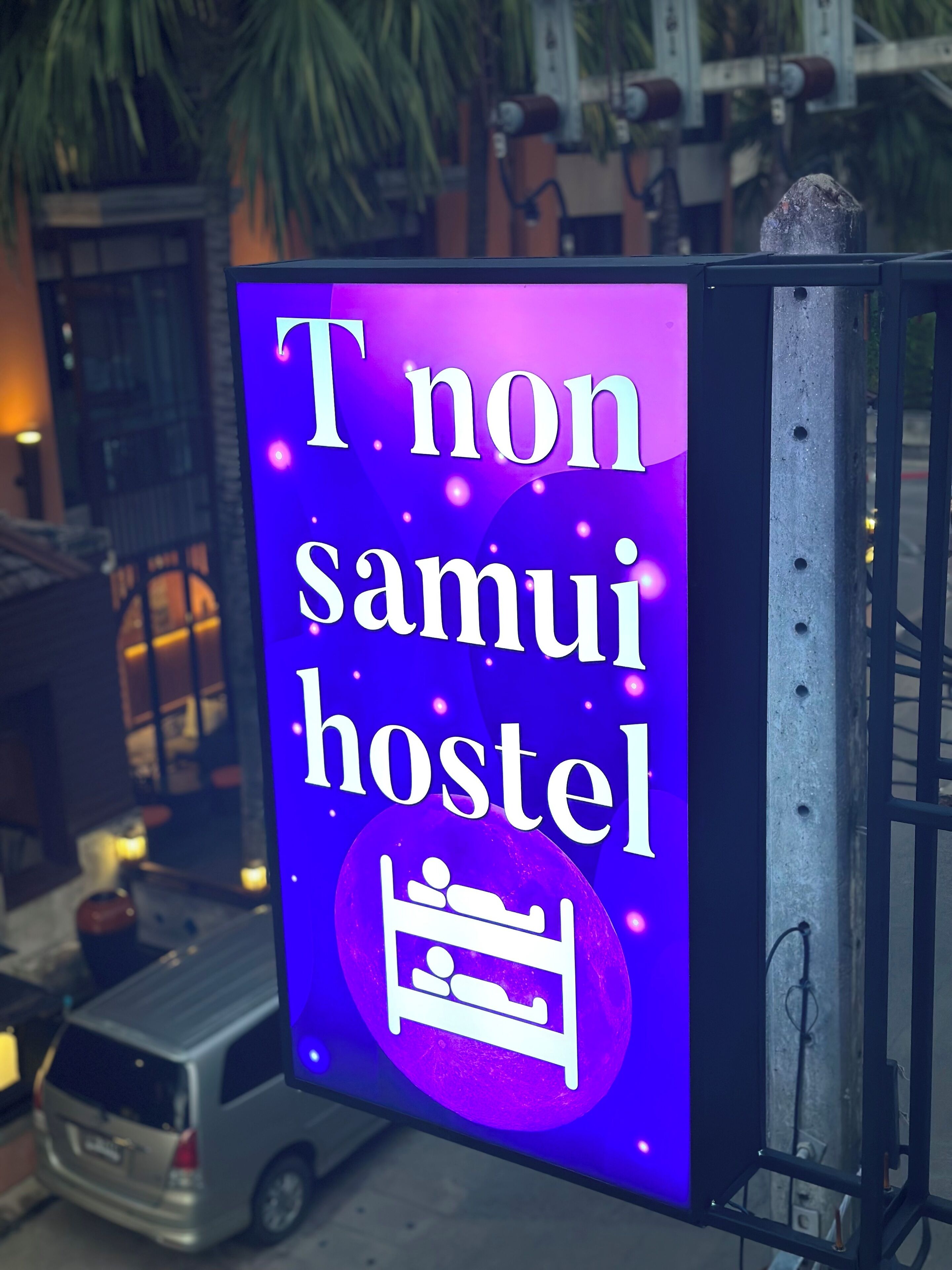 A neon sign with the text "T non samui hostel" and a bed icon, indicating a hostel accommodation.