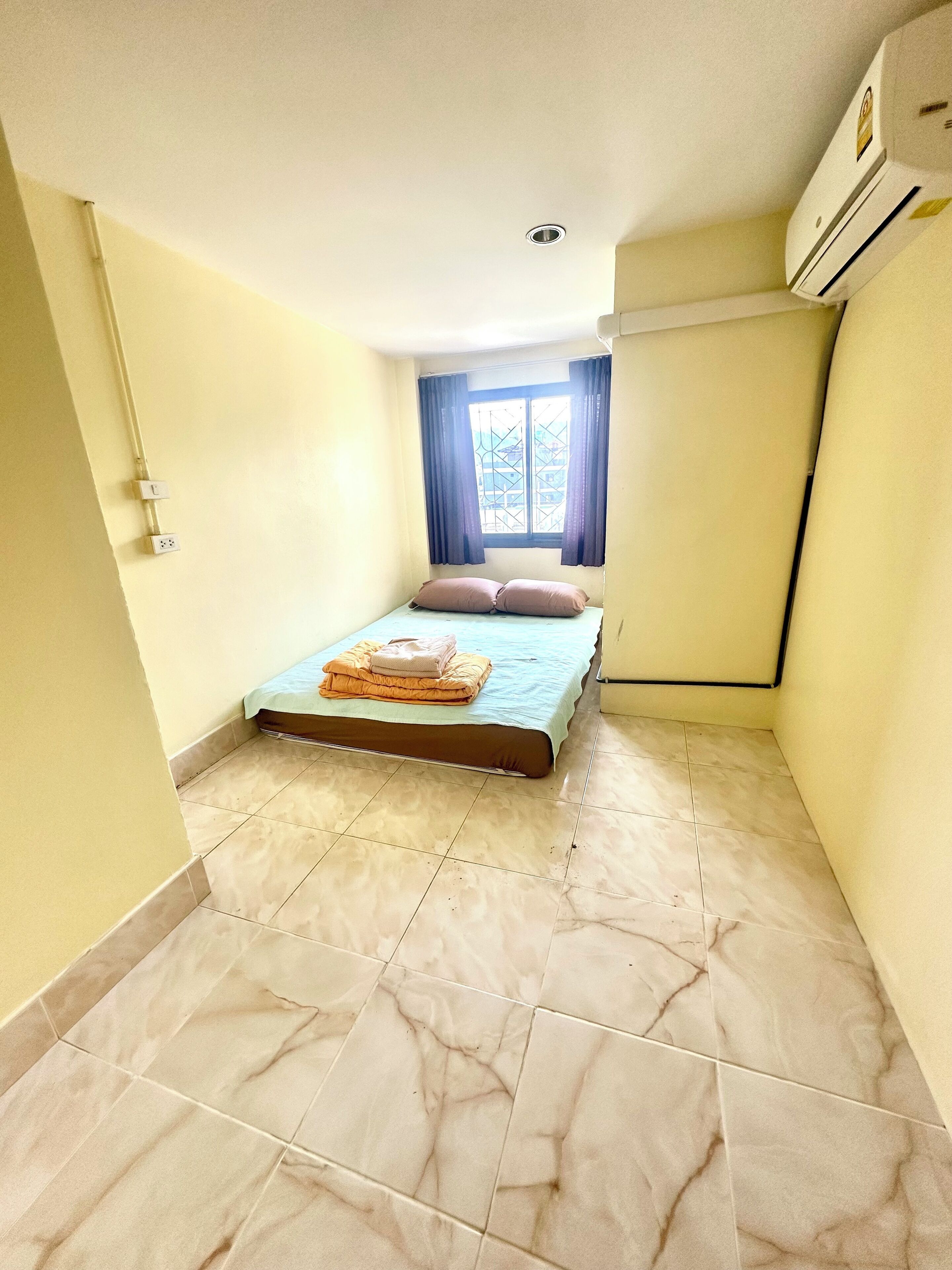 A small, clean room with a bed, a window with curtains, and an air conditioning unit.