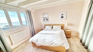 Une chambre à coucher avec un grand lit, une tête de lit en bois, deux tables de chevet avec des lampes, une vue sur la mer par la fenêtre et un radiateur au mur.
