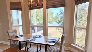 A dining area with a wooden table, four chairs, and a bench. There are large windows with blinds, and a chandelier hangs above the table.