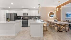 A modern kitchen with white cabinetry, a central island, stainless steel appliances, and a dining area with a round mirror and pendant lights.