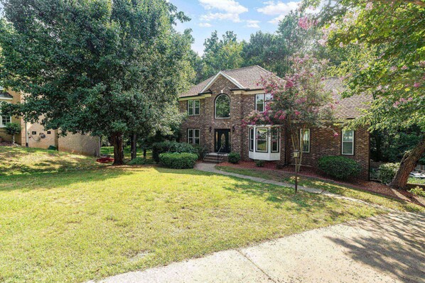A two-story brick house with a front porch, surrounded by a well-maintained lawn and mature trees. - Spacious Mid Term Rental! University City Home with Game Room, Huge Fenced Yard, Pets OK +20% Off! (Charlotte)