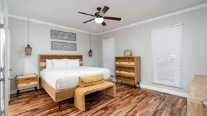 Ein Schlafzimmer mit einem hölzernen Bett, einem Deckenventilator, einem Nachttisch, einer Bank, einer Kommode und einem Fenster mit Jalousien.