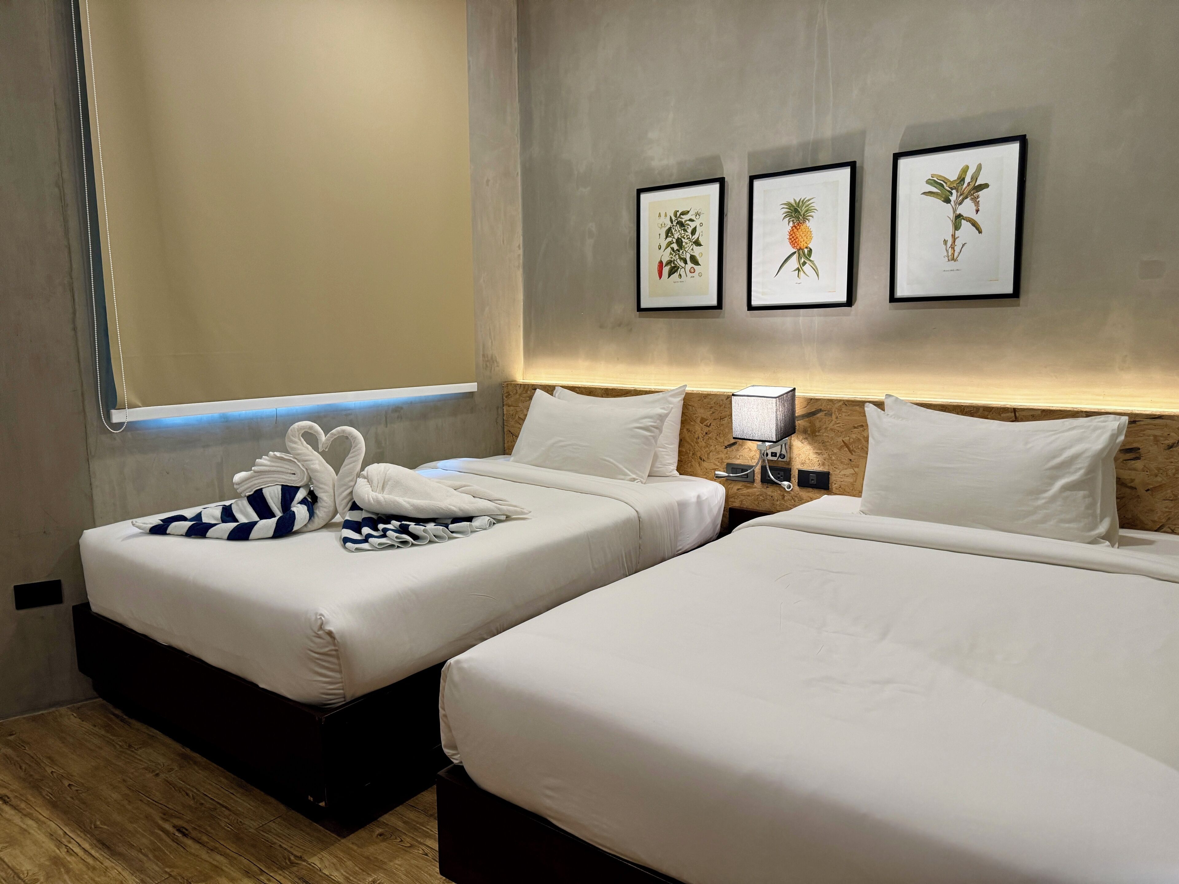 A hotel room with two beds, a nightstand, a wall-mounted lamp, and framed botanical illustrations.