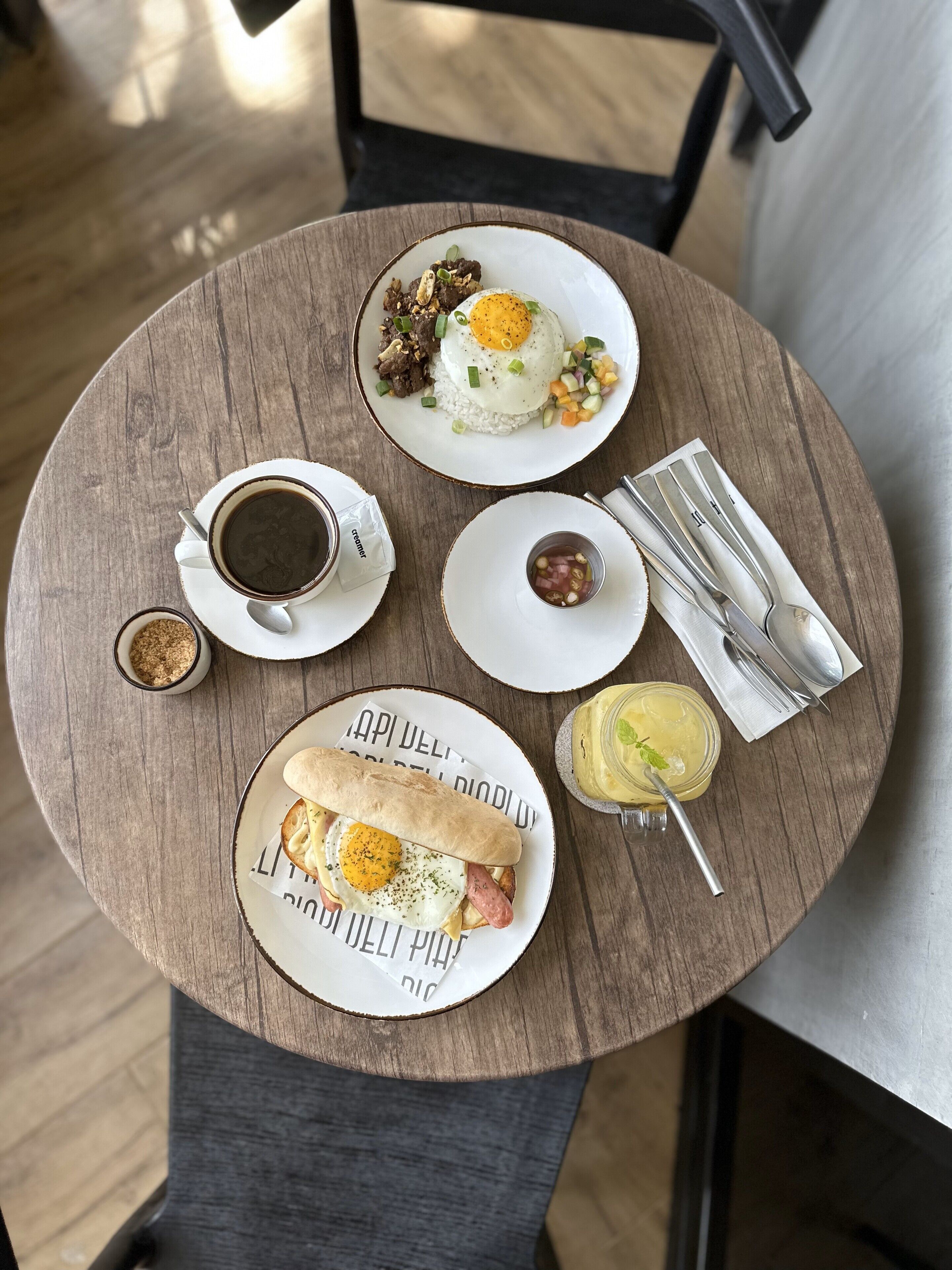 Una mesa redonda de madera con una comida que incluye arroz, huevo, vegetales, una taza de café, un recipiente pequeño con aderezos, un sándwich y una bebida.